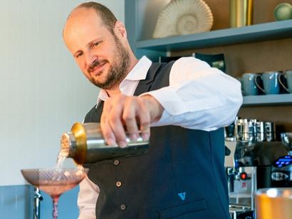 A man wearing a shirt and waistcost is seen pouring a beverage into a cocktail glass.